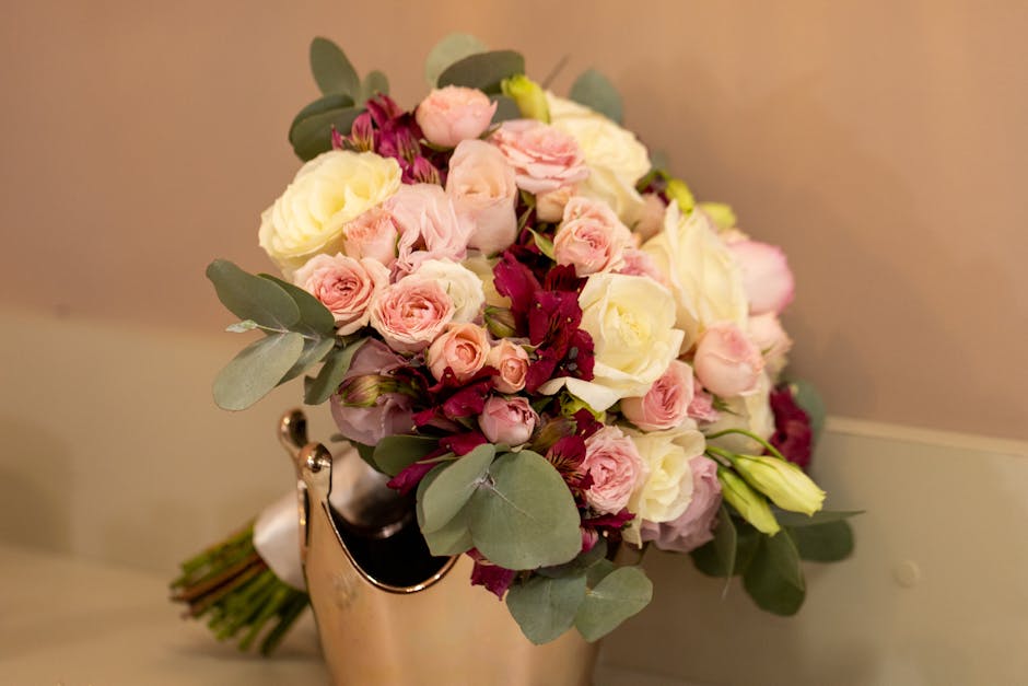 A floral bouquet arrangement resting on a reflective gold vase featuring a mix of fresh flowers including soft pink roses, white roses, pale pink lisianthus, and deep burgundy blooms, accented with lu