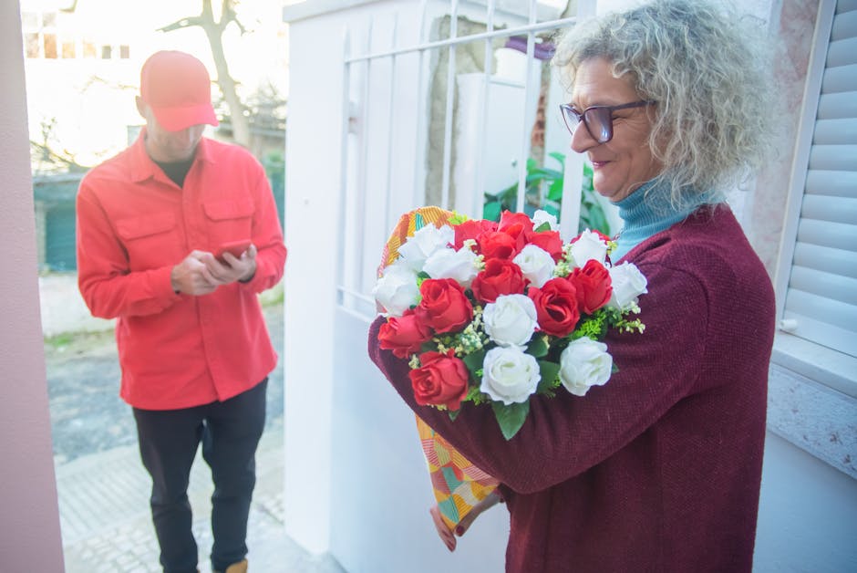 A woman with curly gray hair and glasses is holding a vibrant floral gift consisting of red and white roses, arranged in a bouquet with green foliage. She is wearing a maroon sweater and a blue turtle