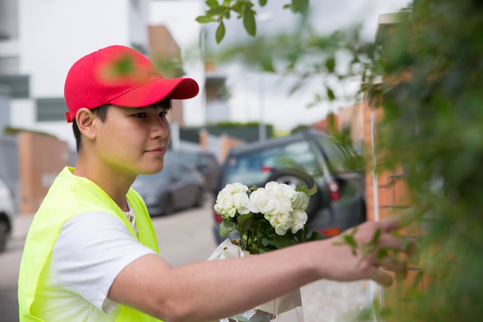 A young male florist wearing a red cap and a high-visibility yellow vest is carefully arranging a bouquet of white hydrangeas with lush green leaves. He is outdoors, standing next to a brick wall part