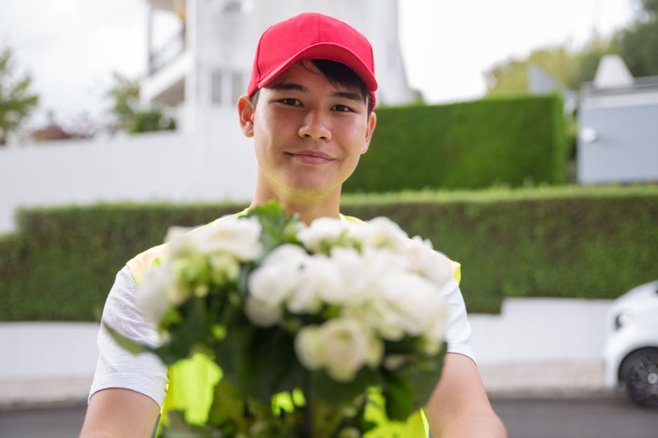 A delivery person wearing a red cap and high visibility vest is smiling warmly while holding a bouquet of fresh white flowers with green foliage. The bouquet features tightly arranged blooms, possibly