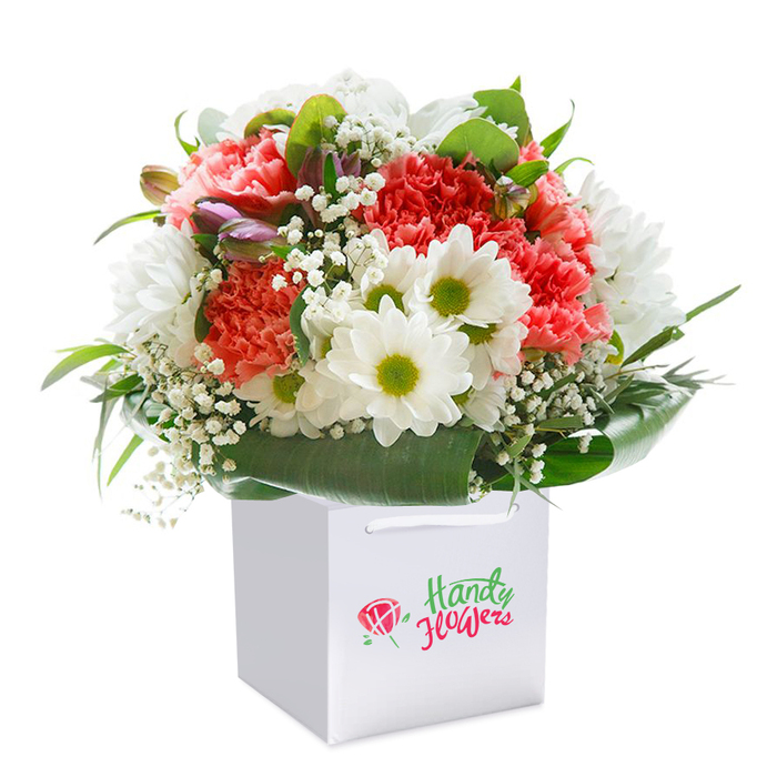 This exquisite bouquet, standing upright in a white square bag with elegant ribbon handles, radiates with the optimism found in Leamouth's lively neighborhoods. The central focus is an ensemble of soft coral-pink and vibrant red carnations, their ruffled blooms arranged to showcase both texture and warmth. Framing the carnations, snowy white daisies with fresh yellow-green centers bring in a crisp, joyful energy, echoing the outdoor spirit of Leamouth's canalside promenades. Airy sprays of baby's breath gently thread between, their cloud-like blossoms imparting softness, while hints of unopened buds brushed with purple add depth and visual intrigue to the composition. The bouquet's foundation is fortified with deep, shining green foliage shaped into a natural wrap-like a floral embrace inspired by the green expanses of nearby East India Dock. Set against a luminous white background, the bouquet appears ready to be presented on a milestone occasion or delivered to brighten a loved one's day along Leamouth Road. Every detail, from the rounded form to the artful colour balance, reflects the expertise and care of local florists dedicated to capturing the beauty of East London in every petal.
