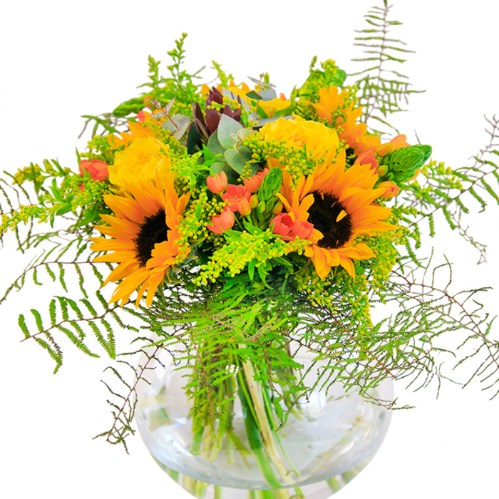 This bouquet makes an immediate statement-sun-soaked exuberance, inspired by the spirited surroundings of Leamouth. The display is dominated by bold, golden sunflowers whose textured faces and yellow-orange petals evoke summer energy, each one standing tall like a beacon beside layers of soft yellow garden roses in full bloom. The dramatic interplay of glossy red Hypericum berries adds a dash of excitement and a nod to the area's vibrant art scene. A rich variety of lush greenery-including drooping fronds of fern, delicate goldenrod blossoms, and slender leafy stems-brings an untamed, organic feel, reminiscent of Leamouth's riverside habitats. Blue-green eucalyptus leaves peek through, gently perfuming the air and introducing a cool undertone to the warmth of yellow. The bouquet sits cozily in a clear, round glass vase, allowing every stem to be admired in its watery chamber-much like the way light reflects off the local waterways. Crafted for celebratory moments or simply to brighten a living space on Orchard Place, this arrangement embodies both the artistry and hospitality for which Leamouth florists are known, offering a sensory experience full of joy, hope, and the memory of sun-filled days along the river.