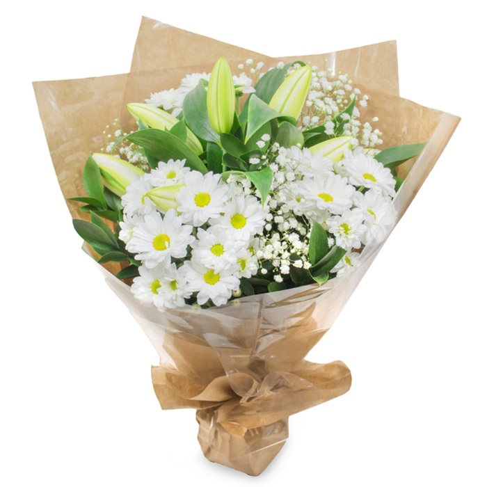This hand-tied bouquet, photographed standing upright against an immaculate white background, showcases a soothing palette of white and green that would feel perfectly at home in a peaceful Leamouth apartment. At its centre, a generous collection of daisy-like white chrysanthemums forms the main body of the design. Each blossom features a ring of fine, snow-white petals surrounding a clear yellow-green disc, providing a subtle focal point that adds energy without overpowering the calm mood. These flowers are set close together, creating a dense, cushioned texture that appears soft to the touch. Piercing gently upwards through the daisies are several slender lily buds, held tightly closed. Their surfaces are smooth and pale green, with creamy hints where the petals almost, but not quite, begin to separate, suggesting fragrance and beauty yet to come. Interspersed among the larger blooms, delicate sprigs of white baby's breath add an airy lace of tiny blossoms, giving the bouquet lightness and an almost floating effect. All of this is supported by lush dark green foliage, its glossy leaves circling the arrangement and adding depth, reminiscent of the greenery that softens the edges of the nearby River Lea. The florist has wrapped the stems carefully in rustic brown kraft paper, the folds and creases giving a handcrafted, natural charm that speaks of care and intention. A clear layer of cellophane envelopes the outer layer, catching glints of light along its faint crinkles and ensuring the flowers are protected during delivery around Leamouth. Gathered and tied securely at the base, the bouquet feels composed and complete, ideal for offering condolences, honouring a cherished memory, or bringing a quiet sense of purity and comfort into a reflective space.