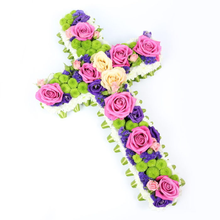Set against a clean, white background, a meticulously crafted floral cross is shown at a slight angle, running from the top left corner towards the bottom right, which lends the design a graceful dynamism. The silhouette of the cross is sharply defined by a continuous border of white spray chrysanthemums, their small, rounded petals overlapping to form a soft but structured frame. Just beyond this snowy trim, a ribbon of pale green material-possibly satin or skillfully folded foliage-wraps around the entire edge, contributing a fresh verdant accent and a polished, professional finish. Within this boundary, the heart of the cross is densely covered in a carefully balanced assortment of blooms. Large roses in vivid fuchsia pink and softer creamy pale yellow provide the main focal points, their open, cupped forms placed thoughtfully along the arms and centre, suggesting points of prayer or reflection. Interspersed among the roses are clusters of bright lime-green button chrysanthemums whose small, spherical heads create a satisfying contrast in both texture and colour, adding a sense of youthfulness and renewal. Smaller flowers in deep purple and violet tones, likely lisianthus or statice, are woven throughout the design, filling gaps and introducing shadowy depth, which tempers the brighter hues and gives the tribute a calm, contemplative air. Every section appears lush and complete, with petals overlapping so that no underlying mechanics are visible. Lit evenly from above, the colours glow softly without glare, making this cross a beautiful option for services in and around Leamouth-whether displayed in a church, a riverside chapel, or a local crematorium-where families seek a floral expression that honours Christian faith while celebrating the vibrancy of the life remembered.